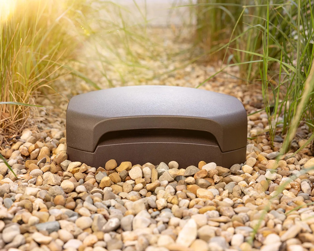A brown, dome-shaped outdoor speaker partially embedded in a bed of small, multicolored pebbles, surrounded by tall green grass with sunlight filtering in from the top left corner.