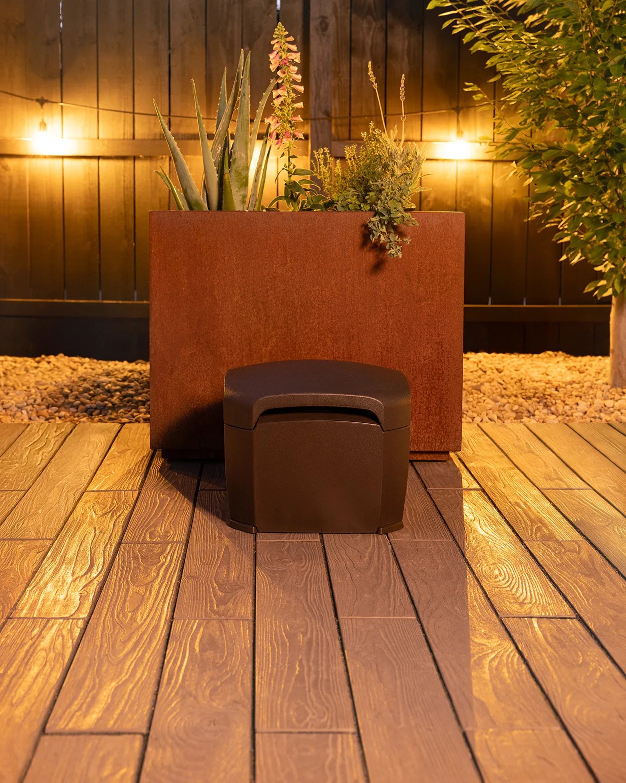 A dark brown outdoor storage box or seat is placed on a wooden deck in front of a large rectangular rust-colored planter filled with various green plants and flowers. The background features a wooden fence with warm string lights, and there are small rocks and a tree on the right side. The scene is illuminated with warm lighting, creating a cozy atmosphere.
