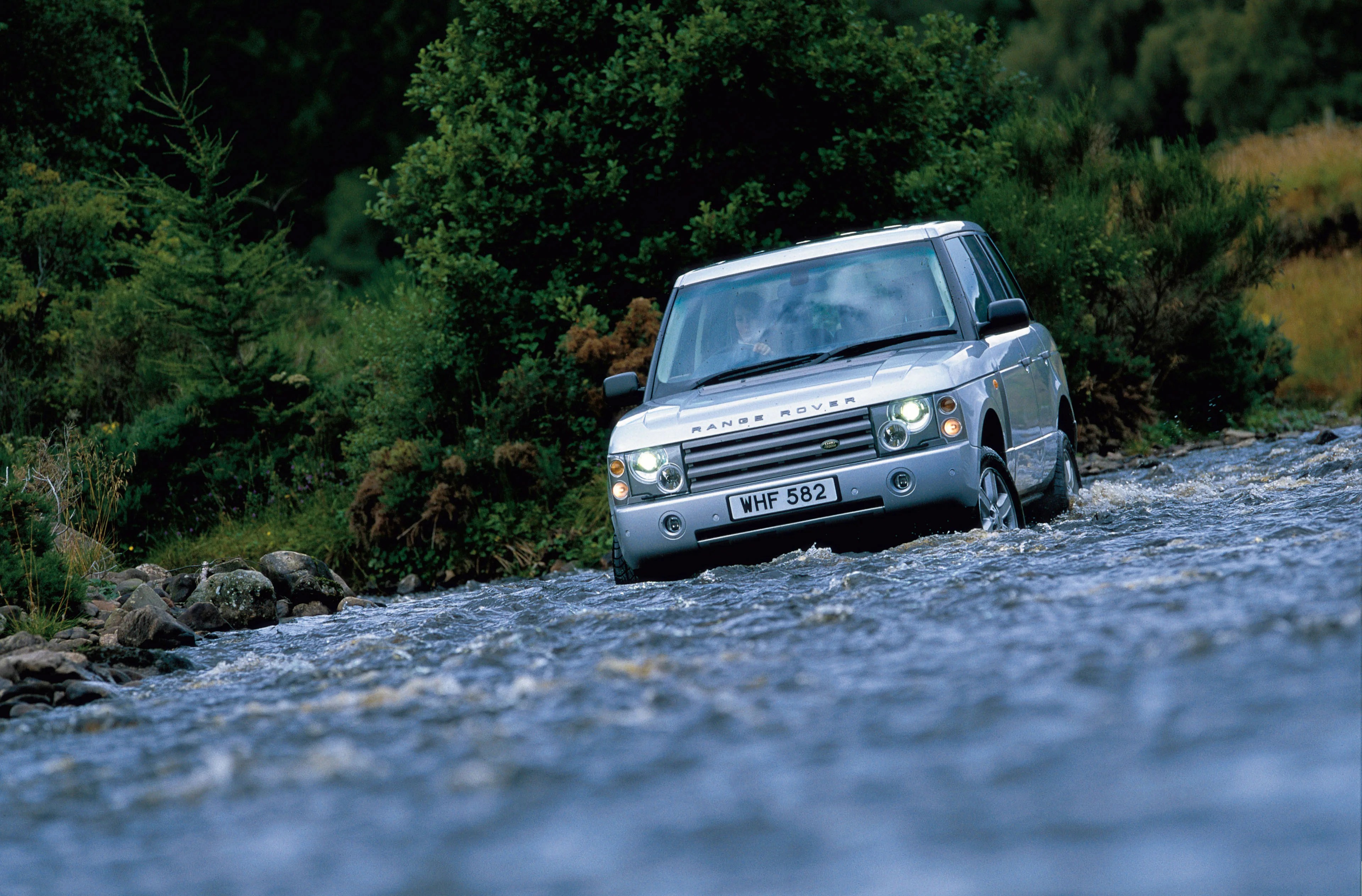 Silver Range Rover driving through a shallow river with water splashing around the tires, surrounded by dense green foliage and rocks along the riverbank.