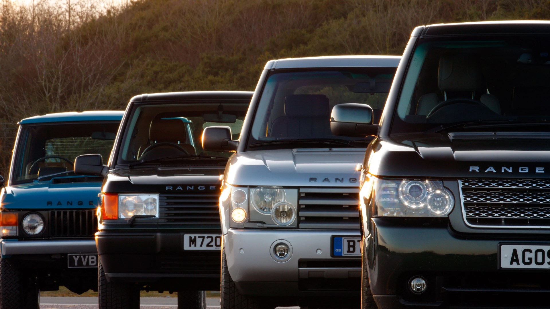 Four Range Rover vehicles are lined up closely side by side, facing forward. The cars vary in color, including black, silver, and blue, and appear to be different model years, showcasing the evolution of the Range Rover design. The background features trees and a softly lit sky, suggesting the photo was taken during sunset or sunrise.