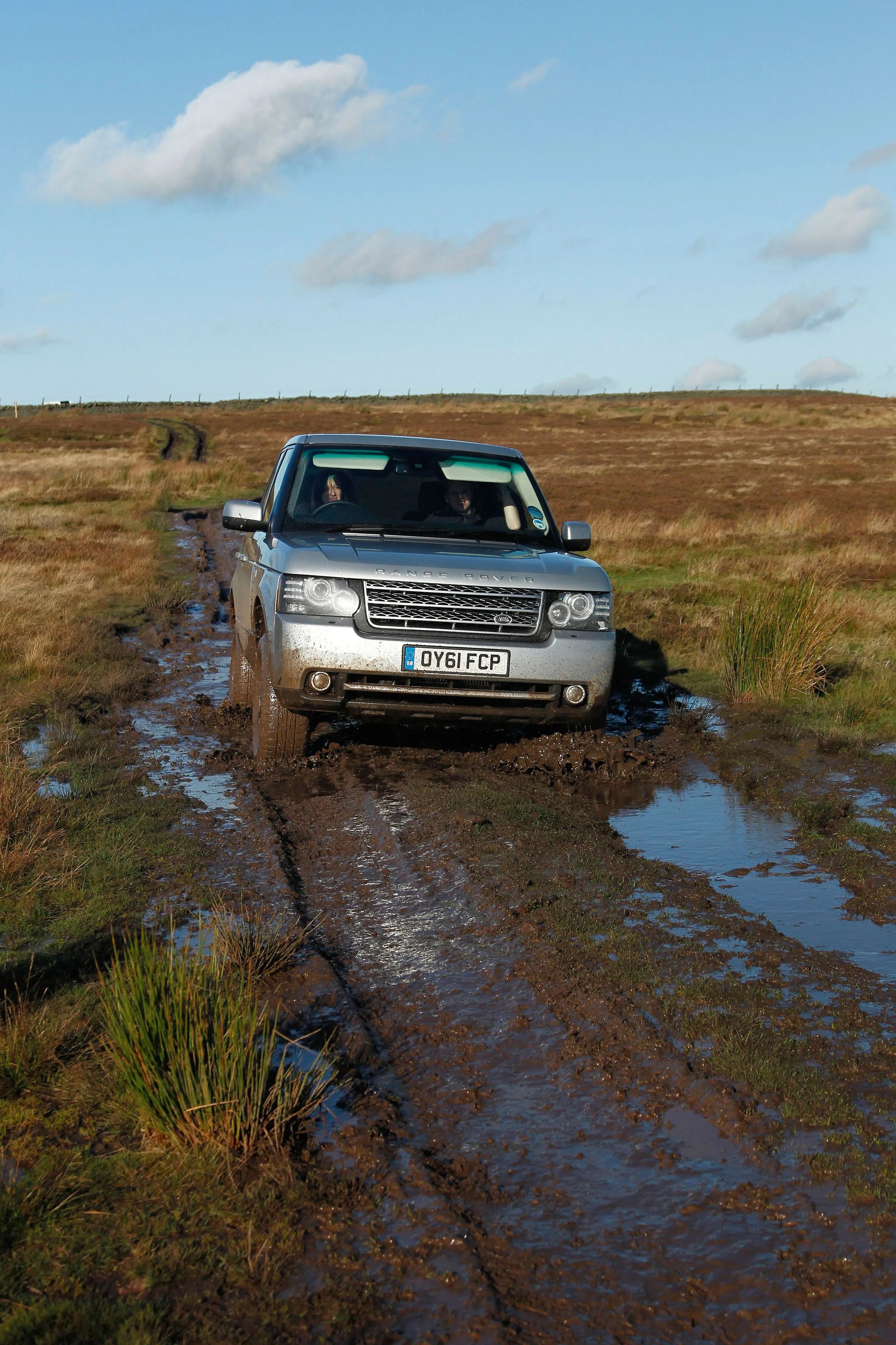 Silver Range Rover driving through a muddy, water-filled dirt track in a grassy, open landscape under a blue sky with scattered clouds. The vehicle's front is splattered with mud, and two people are visible inside.