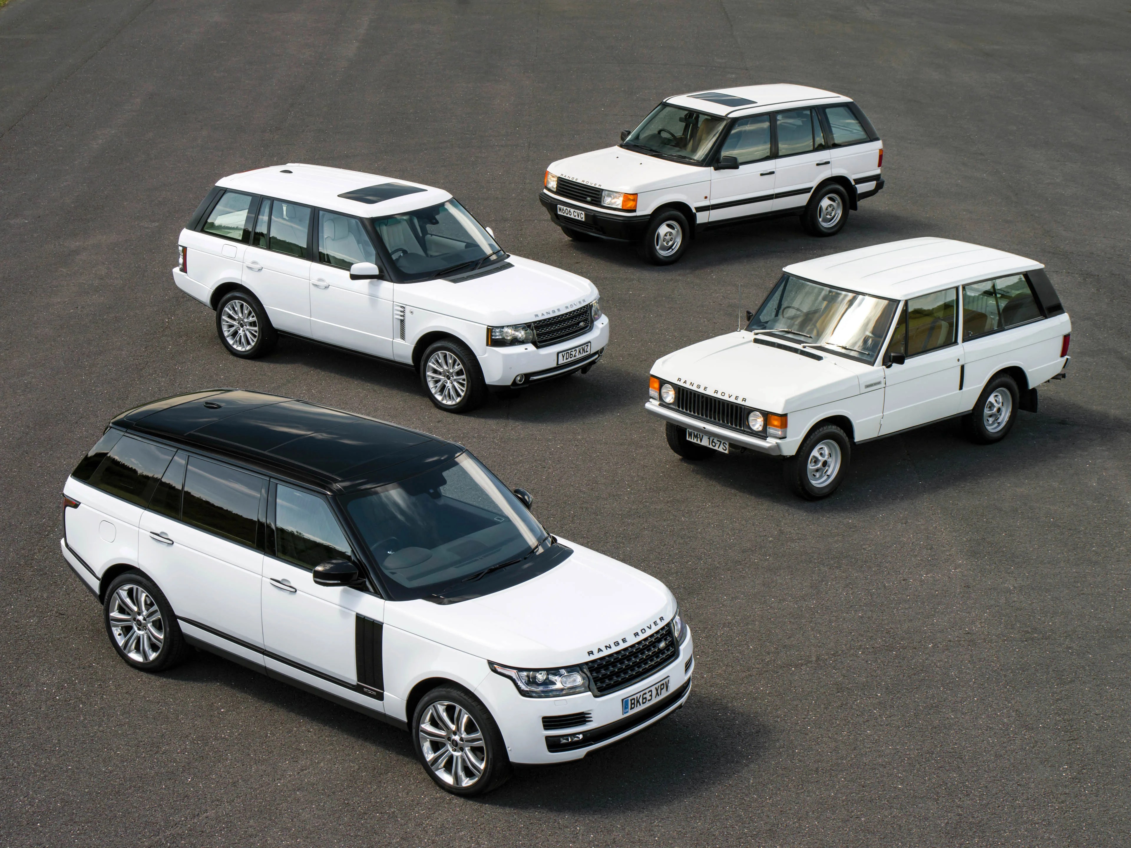 Four white Range Rover SUVs of different generations are parked on an asphalt surface. The vehicles range from a classic early model with a boxy design to more modern versions with sleek lines and updated features. The closest vehicle has a white body with a black roof, while the others are entirely white.