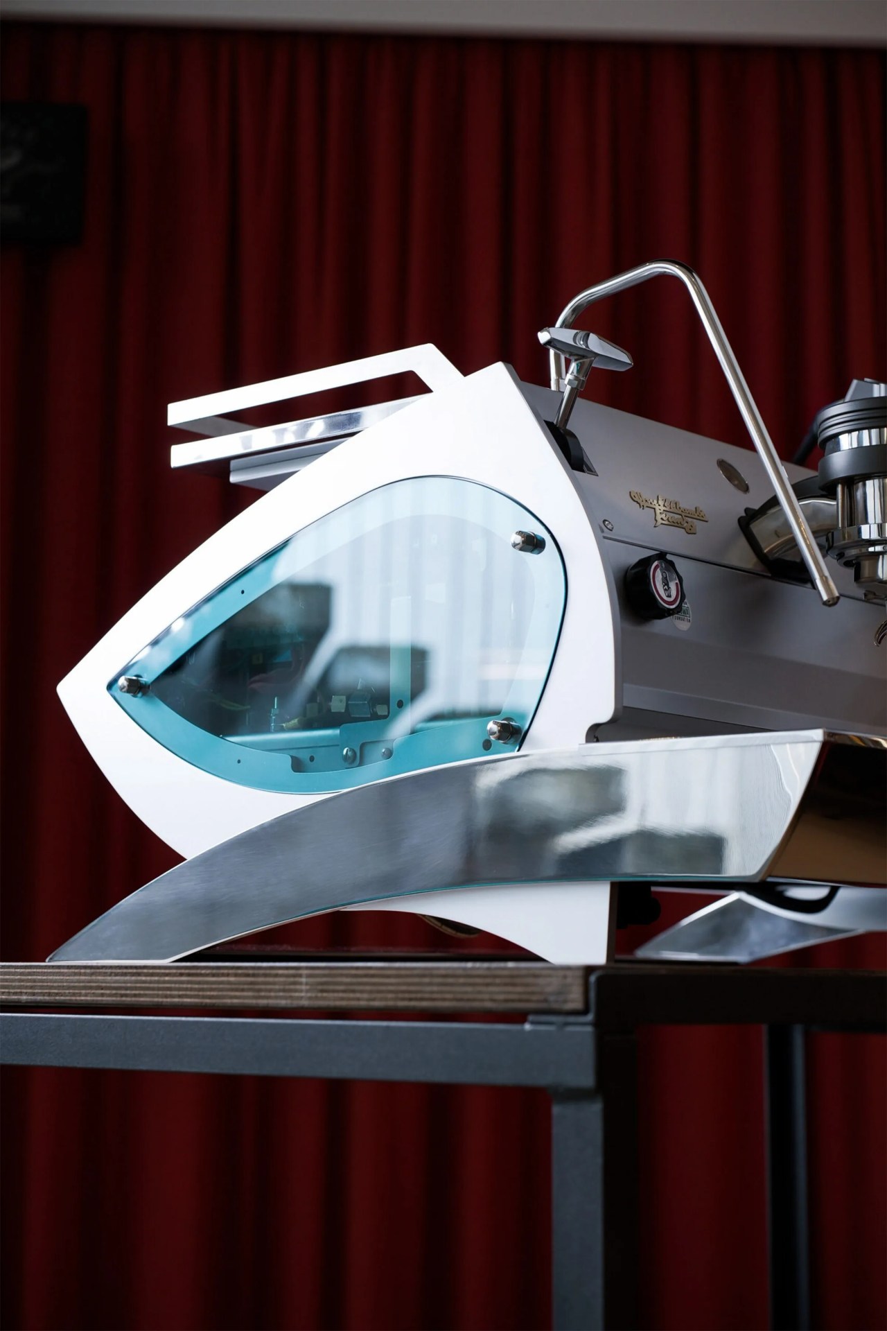 Modern espresso machine with a sleek, angular design featuring a white body, a blue-tinted glass panel on the side, and polished metal accents, placed on a wooden table with a dark metal frame against a backdrop of deep red curtains.
