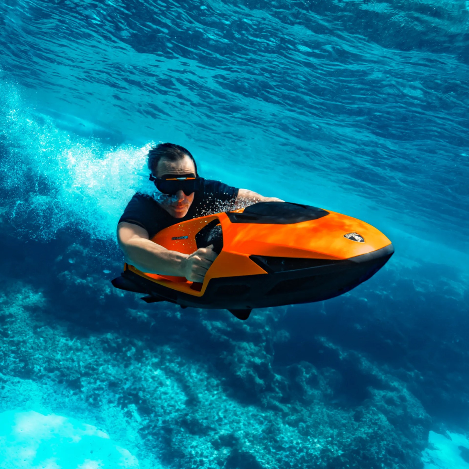 A person wearing a black shirt and black goggles is underwater, riding an orange and black underwater scooter. The water is clear with a blue hue, and the ocean floor with coral is visible beneath. Bubbles trail behind the scooter, indicating movement.