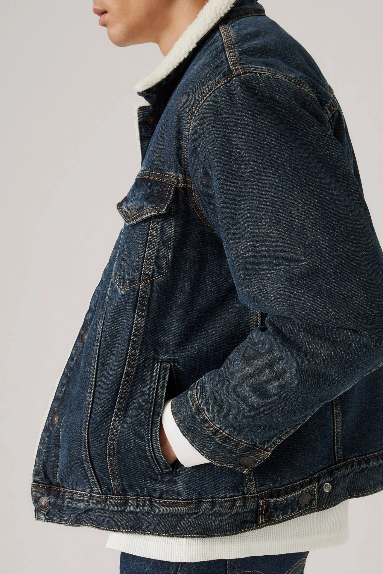 Dark blue denim jacket with a white sherpa collar, featuring chest flap pockets and side pockets, worn over a white ribbed shirt.