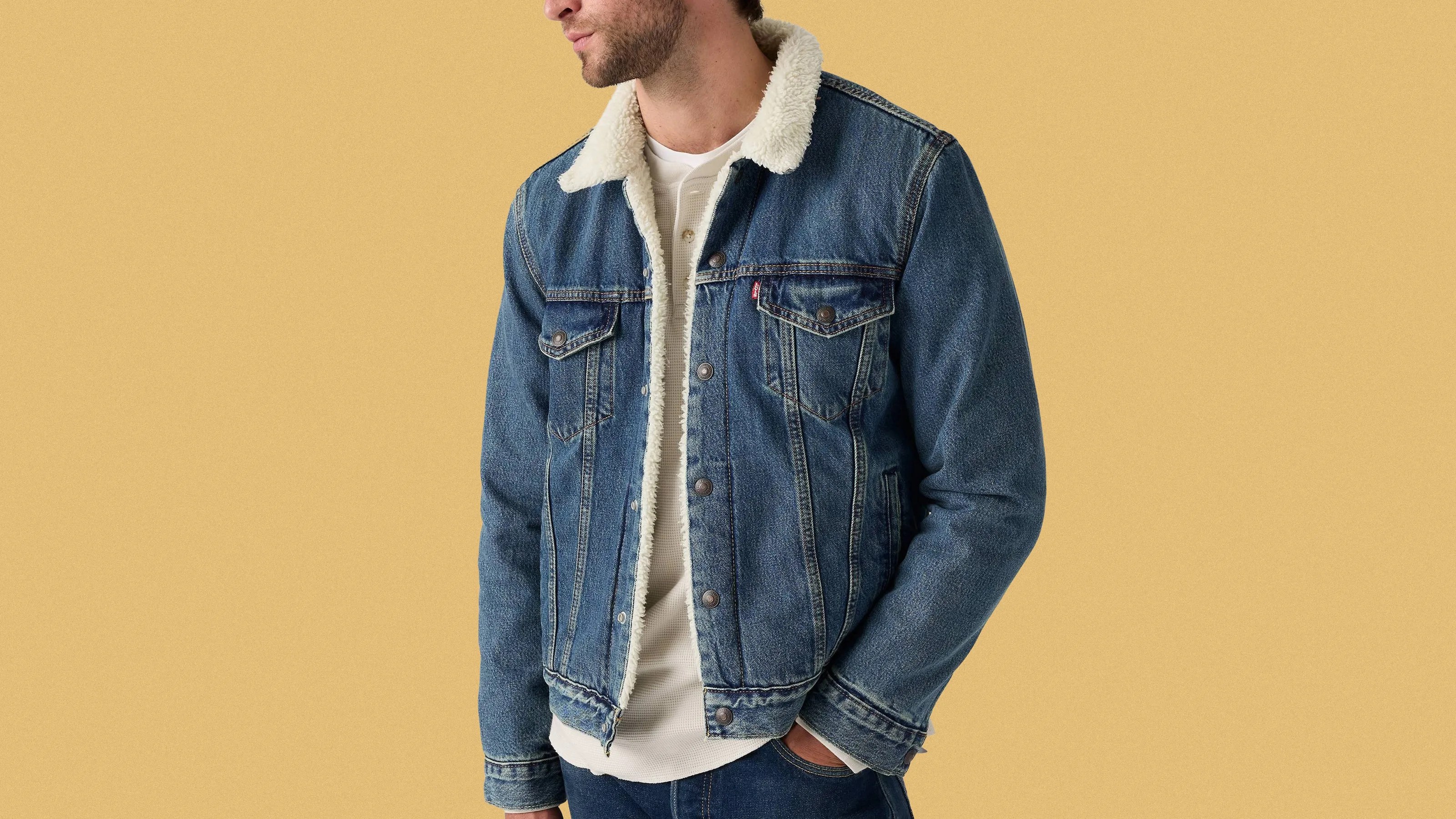 Blue denim jacket with white sherpa lining on the collar and inside, worn over a white henley shirt. The jacket features button closures, two chest pockets with flaps, and side pockets. The person wearing it has one hand in their pocket.
