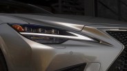 Close-up of a sleek, modern car headlight with a sharp, angular design. The headlight features multiple LED elements and a prominent white LED strip running horizontally. The car's body is metallic silver with smooth, sculpted lines, and part of a black mesh grille is visible on the right side.
