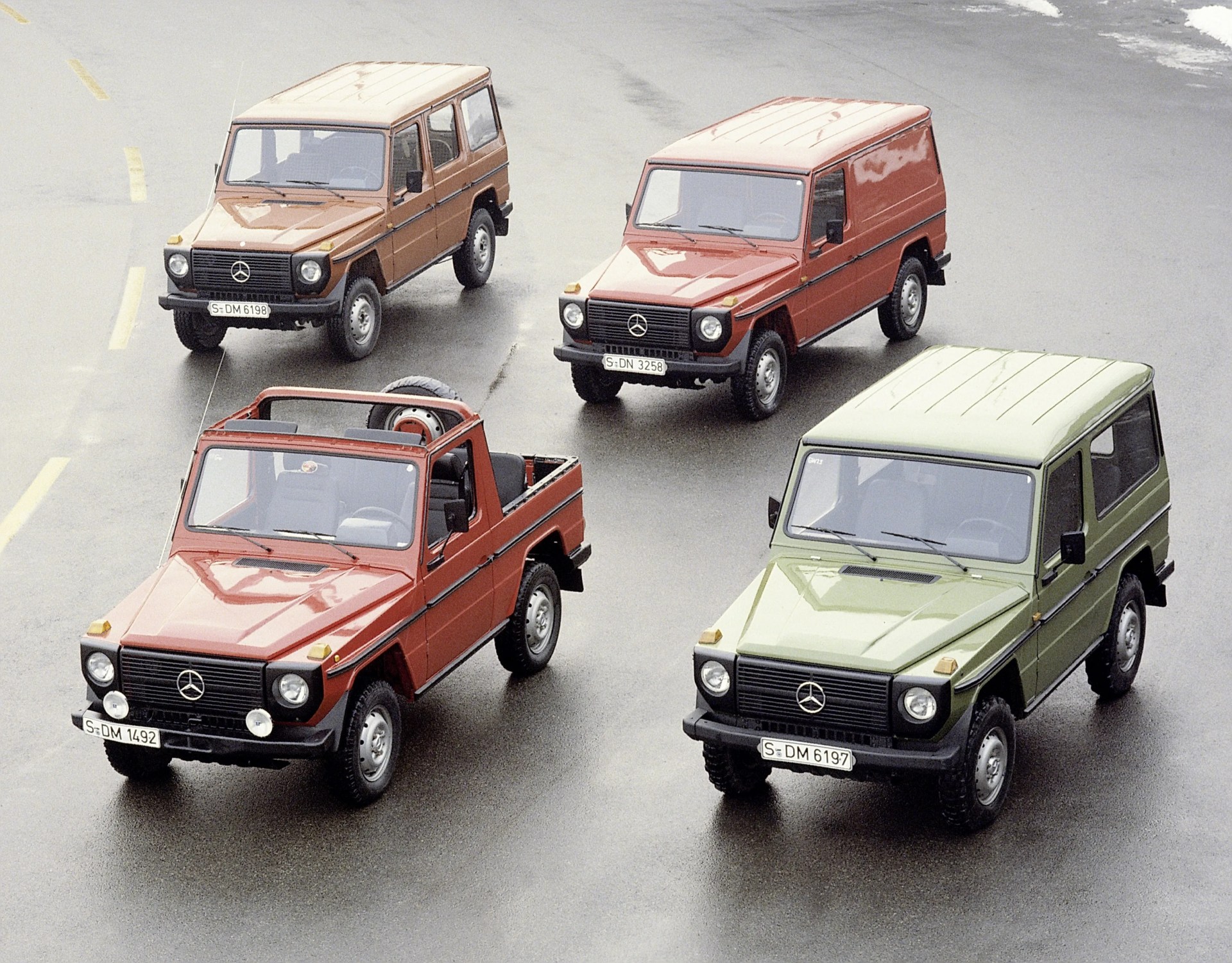 Four classic Mercedes-Benz G-Class vehicles are arranged on a wet asphalt surface. The vehicles include a beige four-door SUV, a red two-door panel van, a red two-door convertible pickup truck with a spare tire in the bed, and a green two-door SUV. All have a boxy design with round headlights and black bumpers.