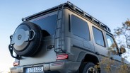 Rear side view of a matte gray Mercedes-Benz G-Class SUV, featuring a spare tire mounted on the back door with a protective cover, a roof rack, and a ladder attached to the rear side. The vehicle has tinted windows and modern LED tail lights. The license plate reads "S G 6325.