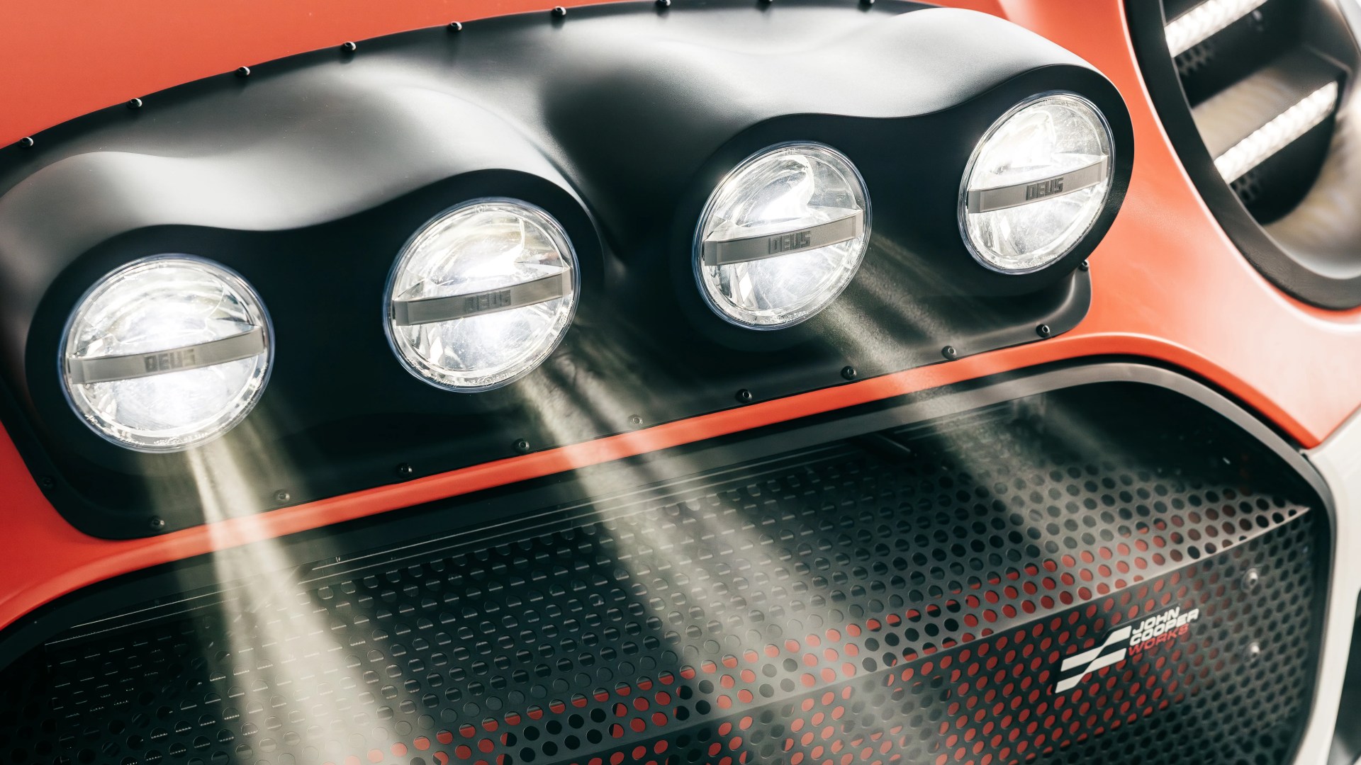 Close-up of four round headlights on a vehicle with a black matte housing and an orange body. Light beams are visible shining from the headlights. Below the lights, there is a perforated grille with a "John Cooper Works" logo.