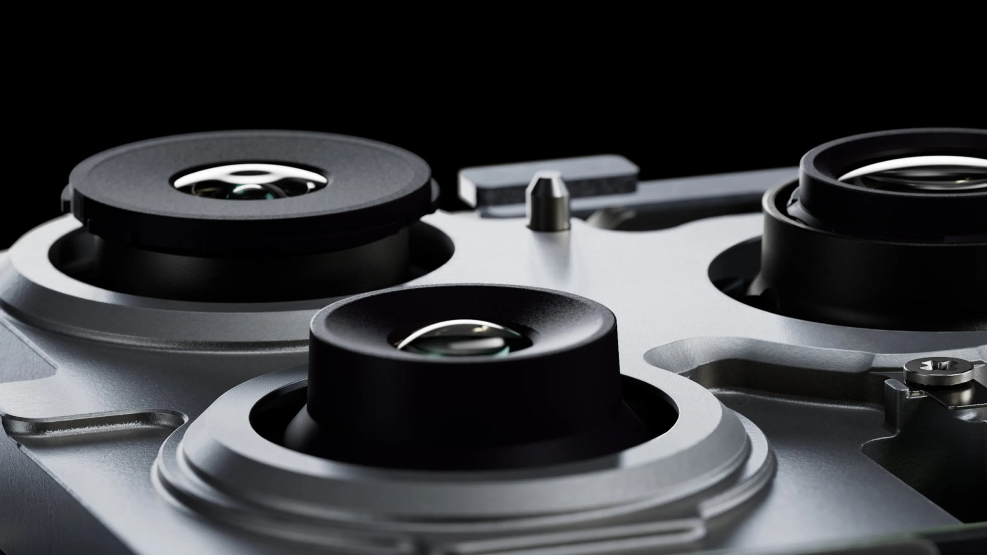 Close-up of a metallic mechanical component featuring three black circular lens-like elements with glass centers, set against a black background. The surface has a smooth, matte finish with visible screws and precise machining details.