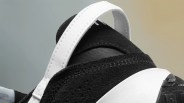 Close-up of the back heel area of a black and white sneaker, featuring a white pull tab with visible stitching and a textured black upper with mesh detail. The midsole is white, and part of the black outsole is visible.