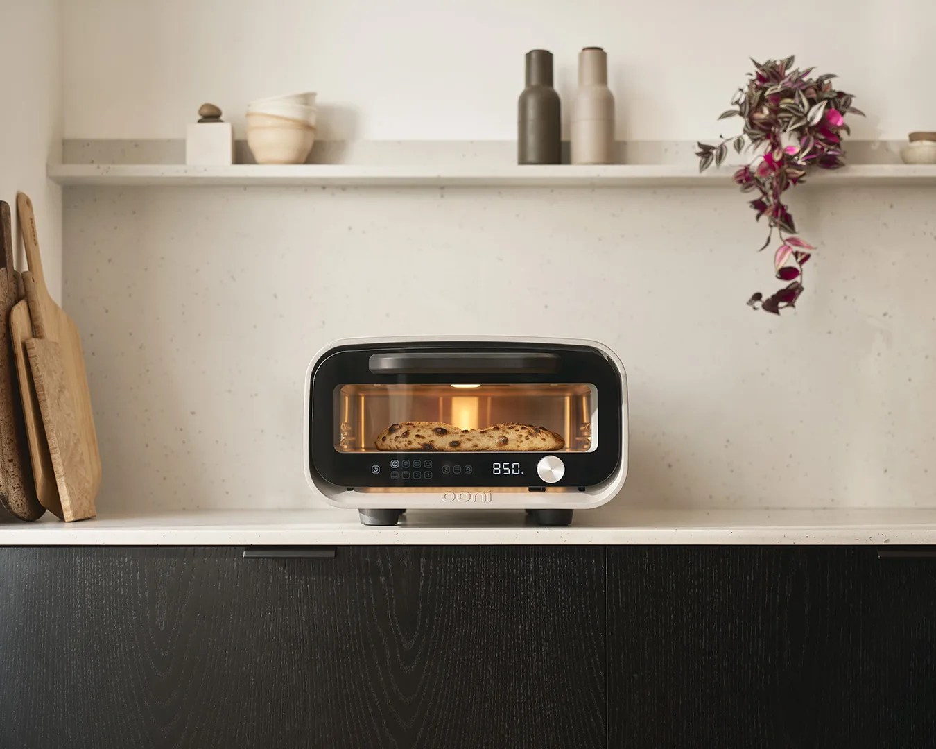 A modern countertop pizza oven with a digital display showing "850" and a silver control knob, baking a pizza inside. The oven is placed on a white countertop with dark wood cabinets below. On the left side of the countertop, there are several wooden cutting boards leaning against the wall. Above the oven, a white floating shelf holds two ceramic bottles, a stack of bowls, a small decorative item, and a trailing plant with purple and green leaves hanging down. The background wall is light-colored with subtle speckles.