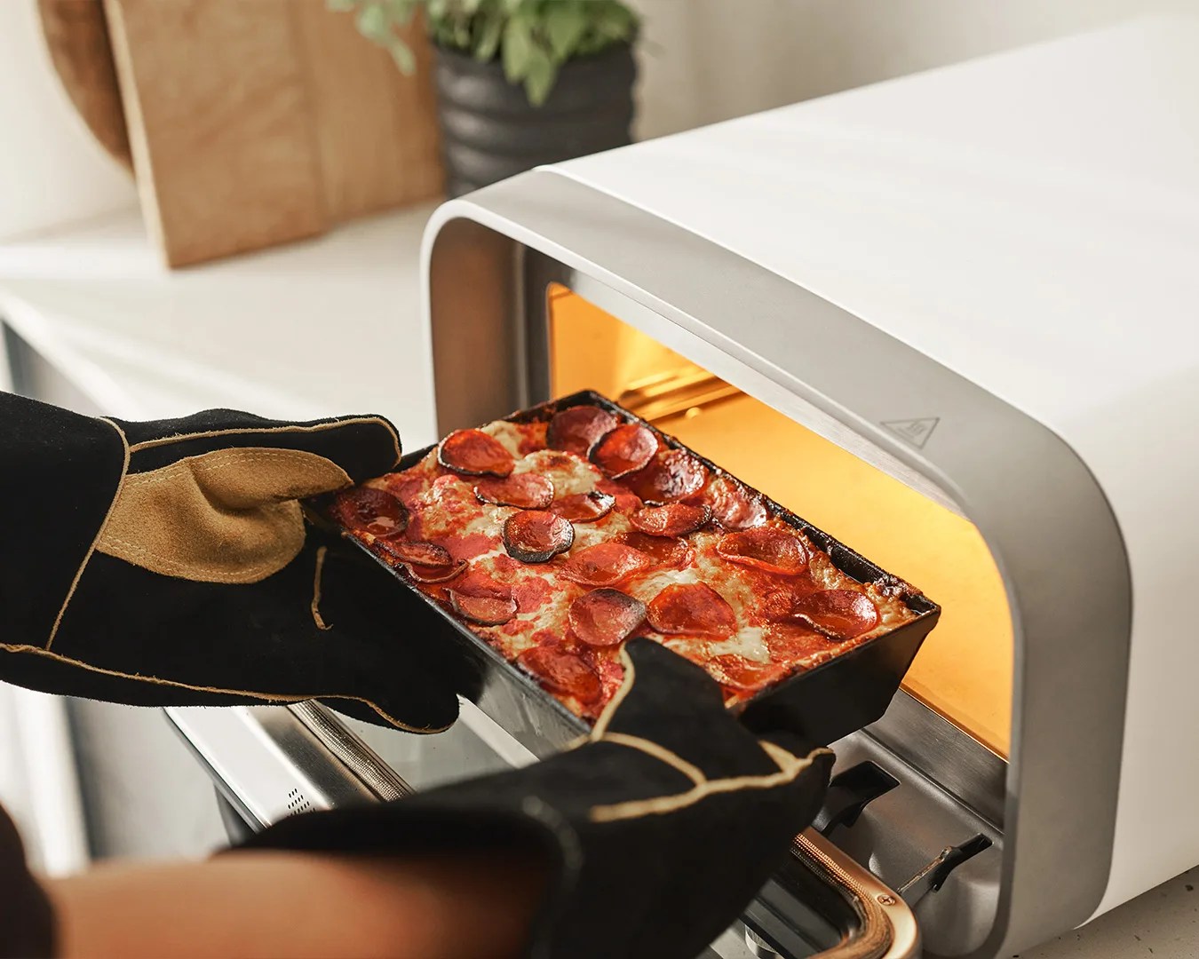 A person wearing black and tan oven mitts is removing a rectangular pepperoni pizza from a small, modern countertop oven with a white and gray exterior. The pizza has melted cheese and evenly distributed pepperoni slices. The oven's interior light is on, illuminating the pizza.