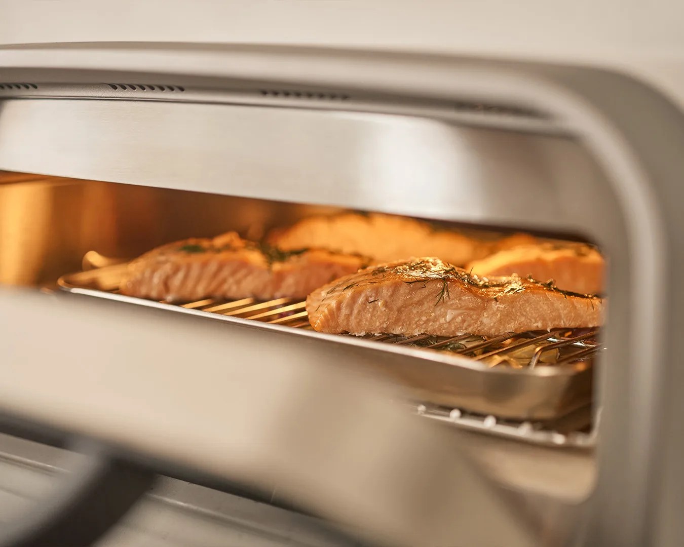 Salmon fillets seasoned with herbs are cooking on a metal rack inside an oven. The oven's interior light casts a warm glow on the fish.
