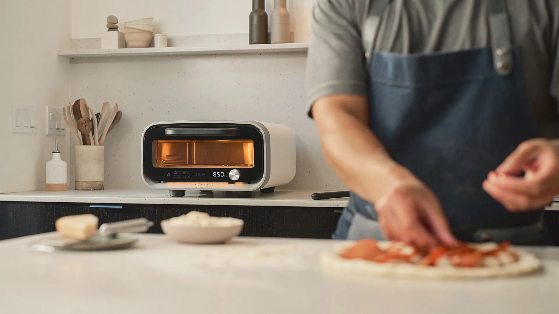 A modern countertop pizza oven with a digital display showing "850" is placed on a kitchen counter. In the foreground, a person wearing a blue apron is preparing a pizza with toppings on a white countertop. Various kitchen utensils and containers are arranged on the counter and a shelf in the background.
