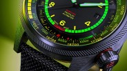 Close-up of an Oris ProPilot Altimeter watch face with a black dial, bright yellow numerals, and green luminous markers. The watch features a black textured bezel with yellow altitude markings in feet, and a black woven strap. The crown has red accents and markings. The background is a gradient green.