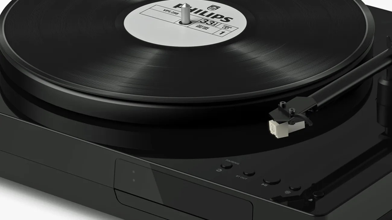 Black turntable with a vinyl record labeled "PHILIPS" on it, featuring a tonearm and cartridge positioned near the edge of the record. The turntable has control buttons including "PHONES," "BT OUT," and play/pause icons on its glossy surface.