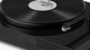 Black turntable with a vinyl record labeled "PHILIPS" on it, featuring a tonearm and cartridge positioned near the edge of the record. The turntable has control buttons including "PHONES," "BT OUT," and play/pause icons on its glossy surface.