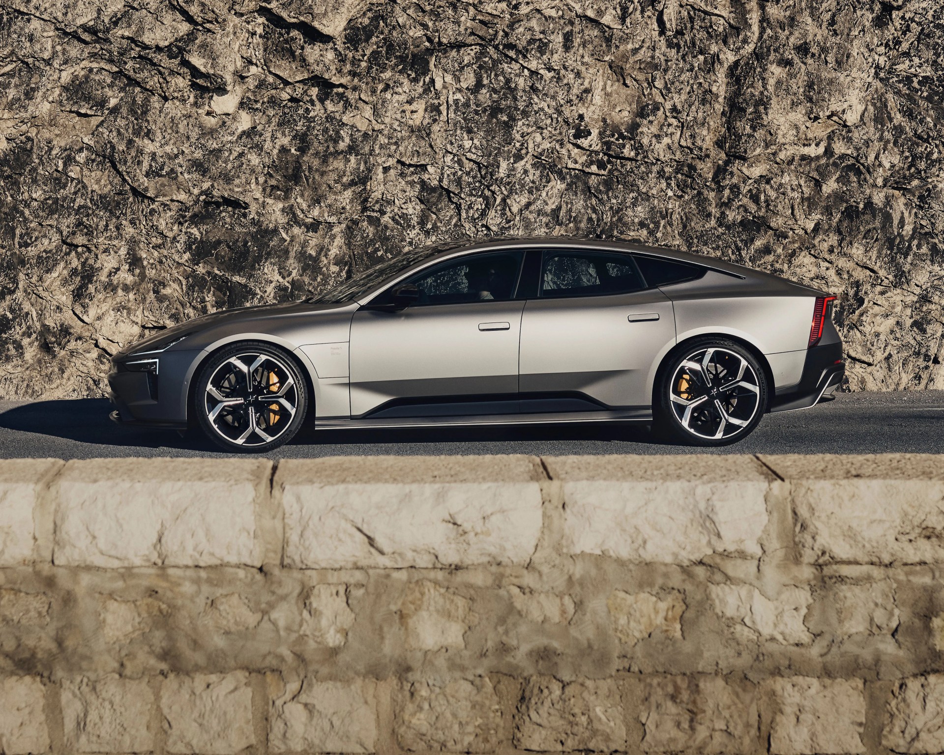 Sleek silver electric sedan with black accents, featuring modern aerodynamic design, large black and silver alloy wheels, and yellow brake calipers, parked on a road with a textured rocky wall in the background.