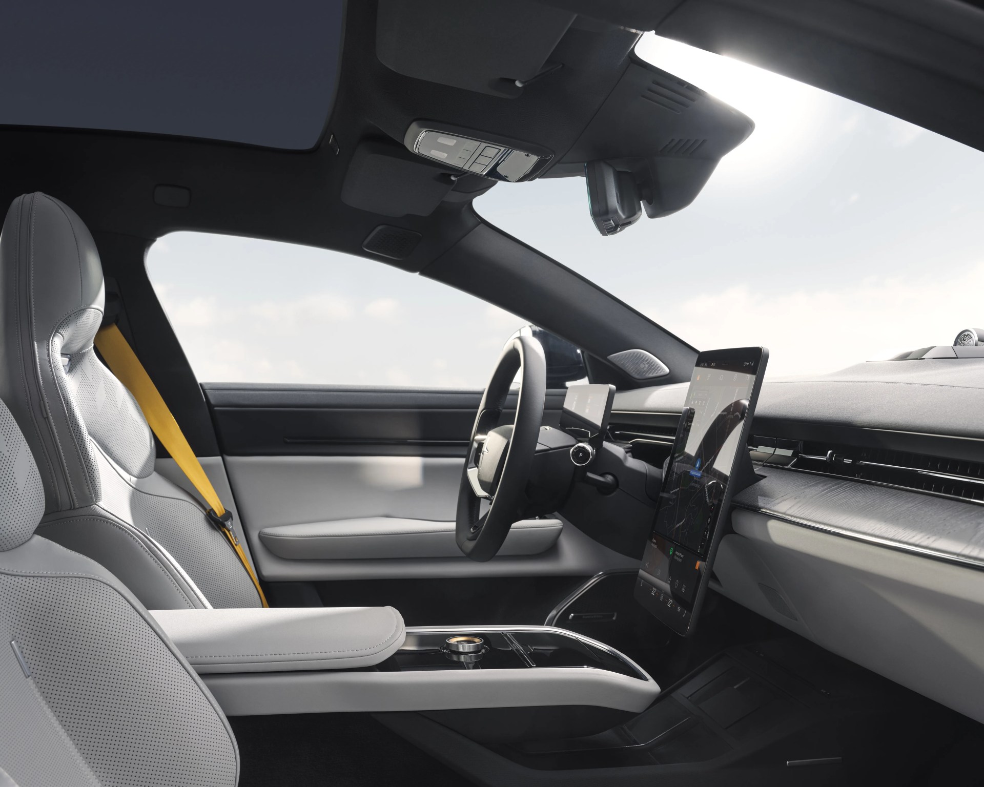 Interior of a modern car featuring light gray perforated leather seats with a yellow seatbelt, a large vertically oriented touchscreen display on the dashboard, a minimalist steering wheel, and a panoramic glass roof. The center console has a sleek design with a rotary control knob and an armrest. The dashboard includes a horizontal air vent and a wood-textured trim.
