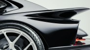 Close-up of the rear section of a sleek black sports car, highlighting the aerodynamic rear wing, part of the rear wheel with a silver rim, and a carbon fiber diffuser. The car's surface is glossy, reflecting light smoothly.