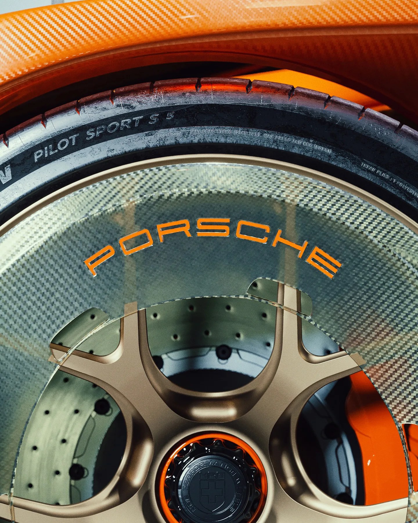 Close-up of a Porsche wheel featuring a carbon fiber cover with the word "PORSCHE" in orange letters, bronze-colored spokes, a black and orange center cap, and a Michelin Pilot Sport 5 tire. The brake disc behind the wheel is visible with drilled holes. The car's body is orange with a carbon fiber texture.