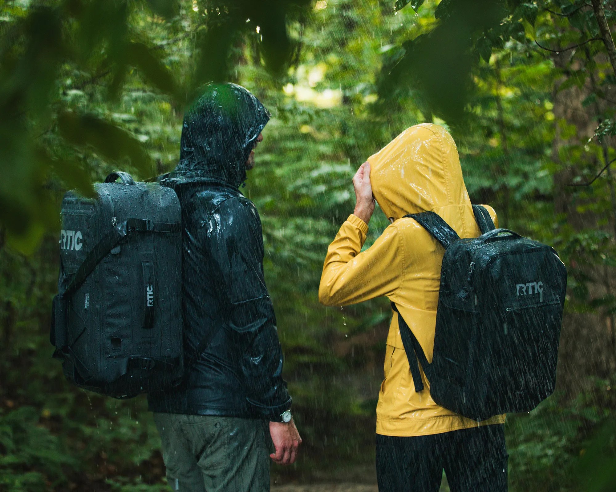 Two people wearing rain jackets and backpacks are standing in a forest during heavy rain. One person is in a black rain jacket with a large black backpack, and the other is in a yellow rain jacket with a smaller black backpack. Both backpacks have the "RTIC" logo. The person in the yellow jacket is adjusting their hood.