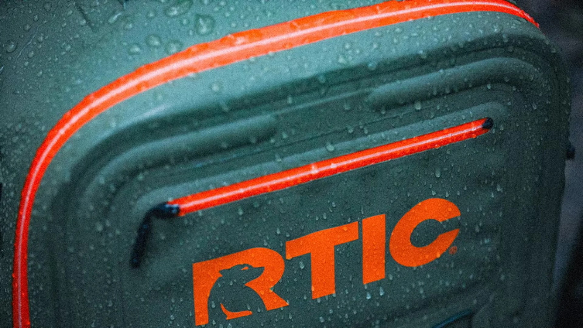 Close-up of a green RTIC cooler or bag with orange zippers and logo, covered in water droplets.