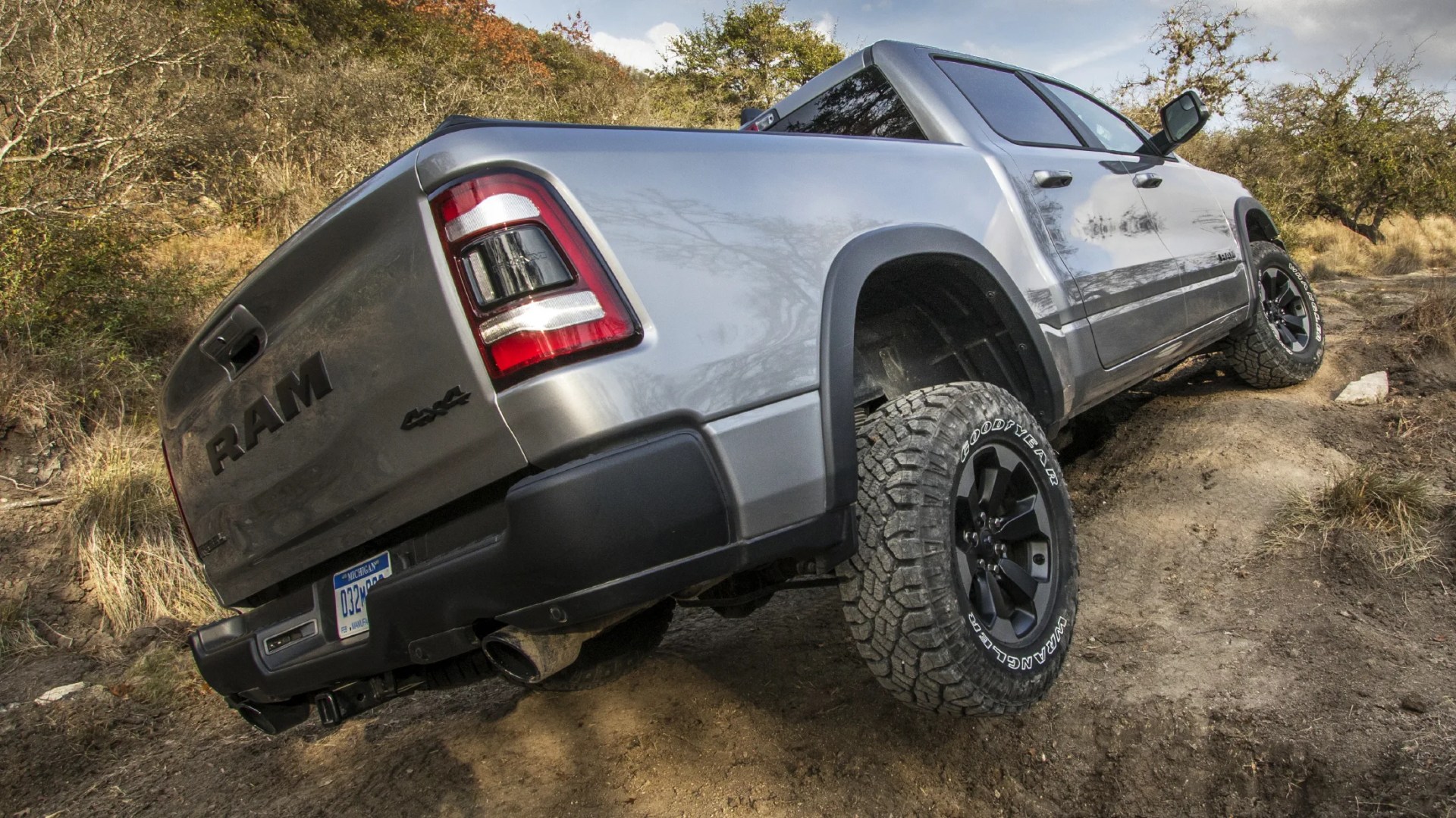 Silver RAM 4x4 pickup truck with black wheels and Goodyear Wrangler tires driving off-road on a dirt trail surrounded by dry grass and bushes.
