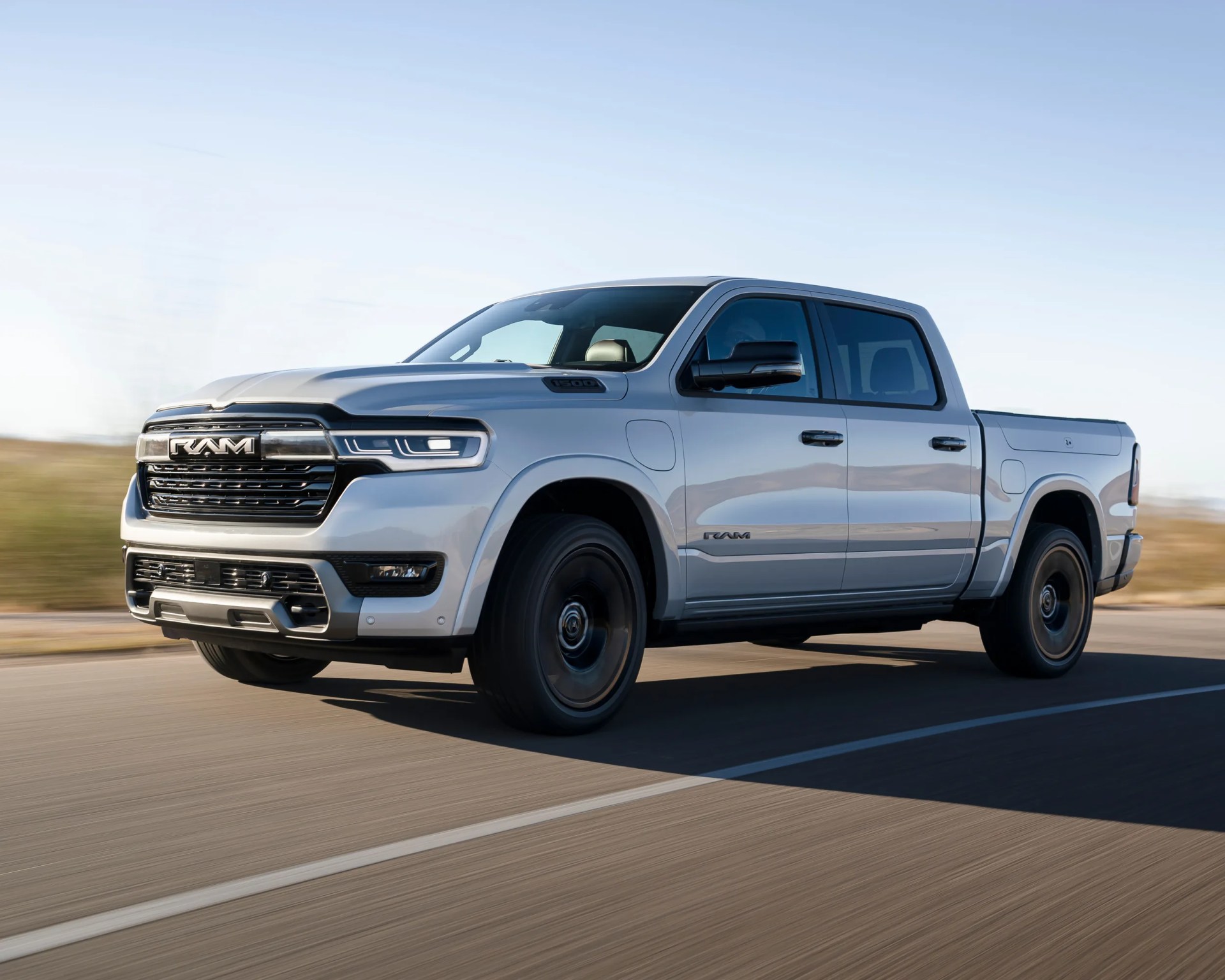 Silver Ram 1500 pickup truck driving on a paved road with a clear sky background. The truck has a crew cab, black wheels, and a prominent front grille with the RAM logo.