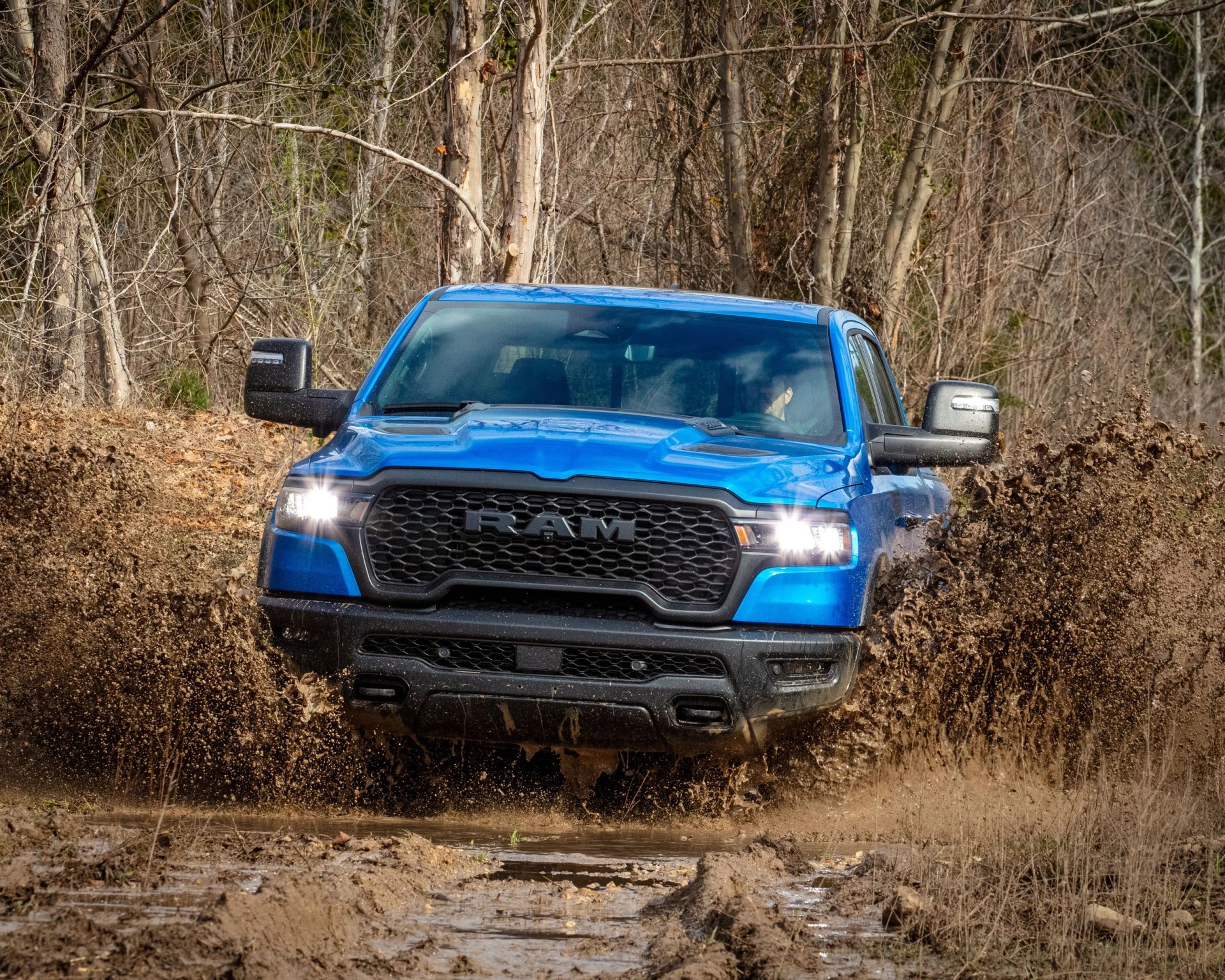 Blue RAM pickup truck driving through a muddy forest trail, splashing mud on both sides, with headlights on and a driver visible inside.
