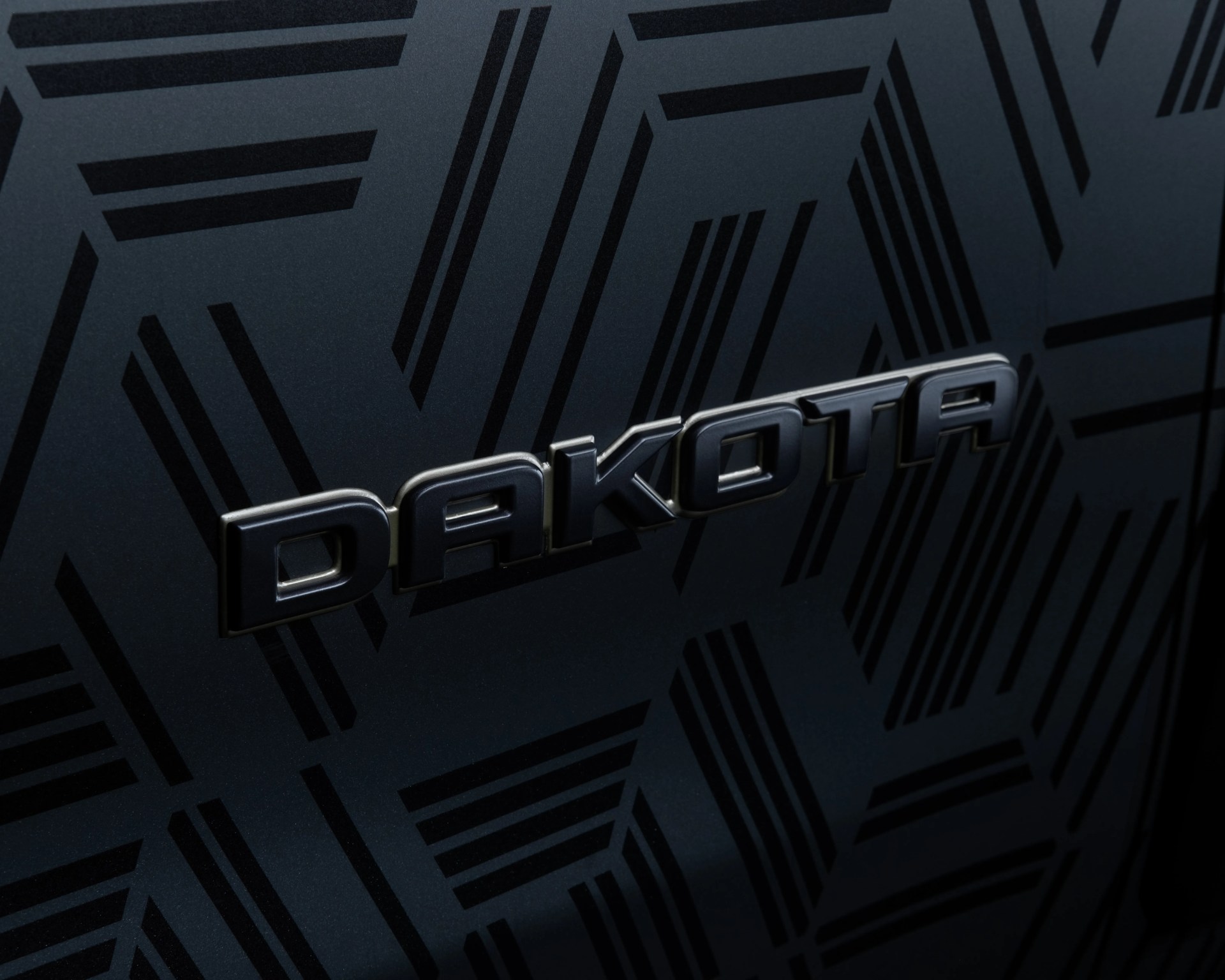 Glossy black "DAKOTA" emblem on a dark surface with a geometric pattern of black lines and shapes.