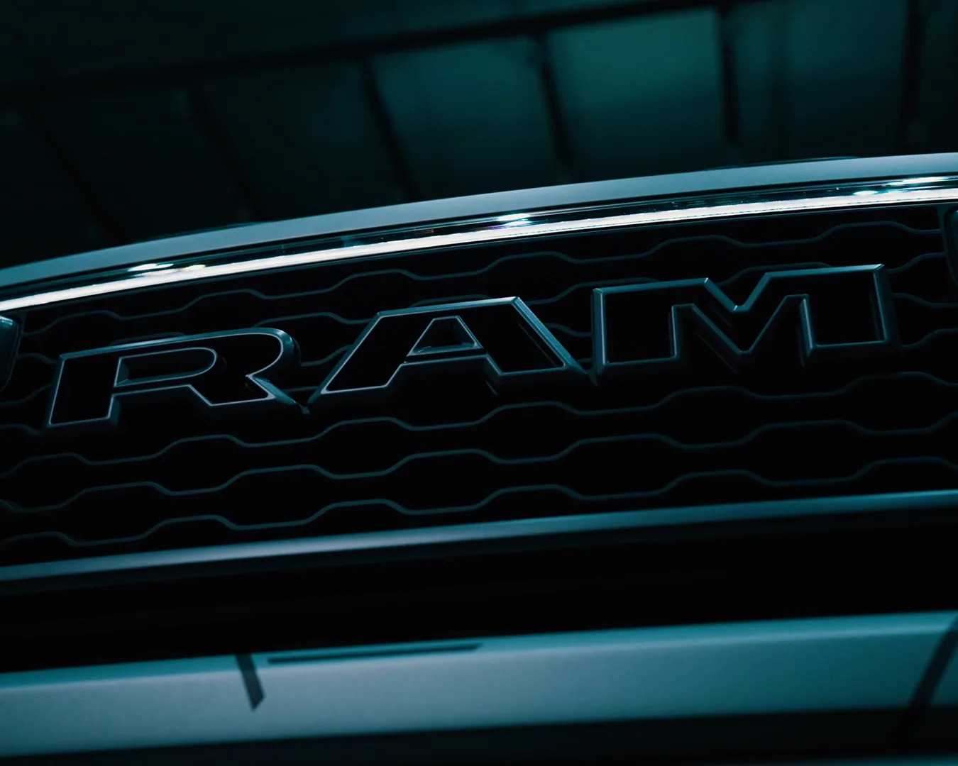 Close-up of a RAM truck grille with the RAM logo prominently displayed in dark, moody lighting. The grille features a wavy pattern behind the bold, raised letters.