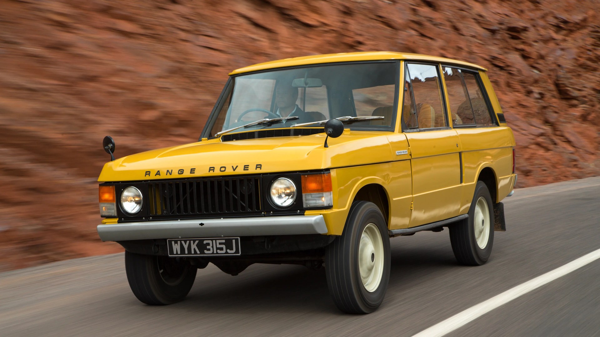 Yellow classic Range Rover SUV driving on a road with a rocky, reddish-brown background, featuring round headlights, black grille, and white wheels.