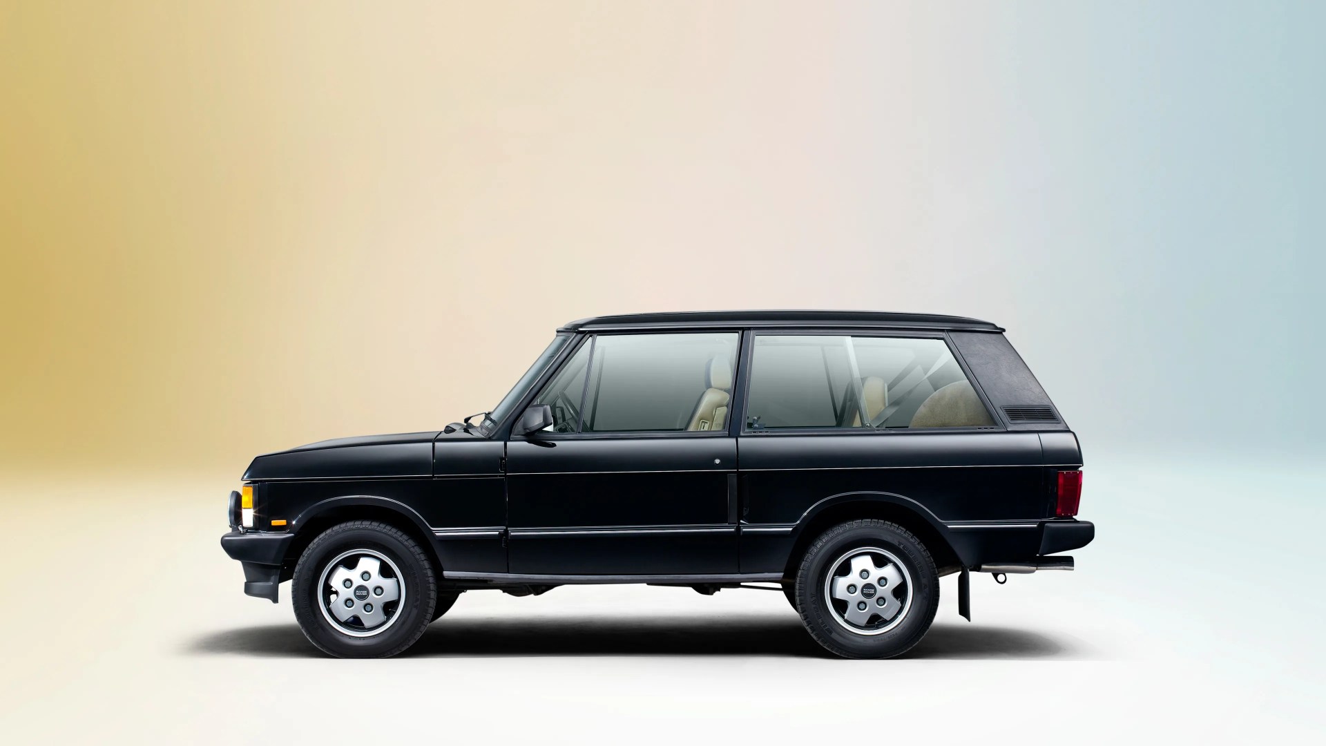 Black classic two-door SUV with beige interior, featuring a boxy design and silver alloy wheels, shown in profile against a gradient background transitioning from warm beige to cool blue.