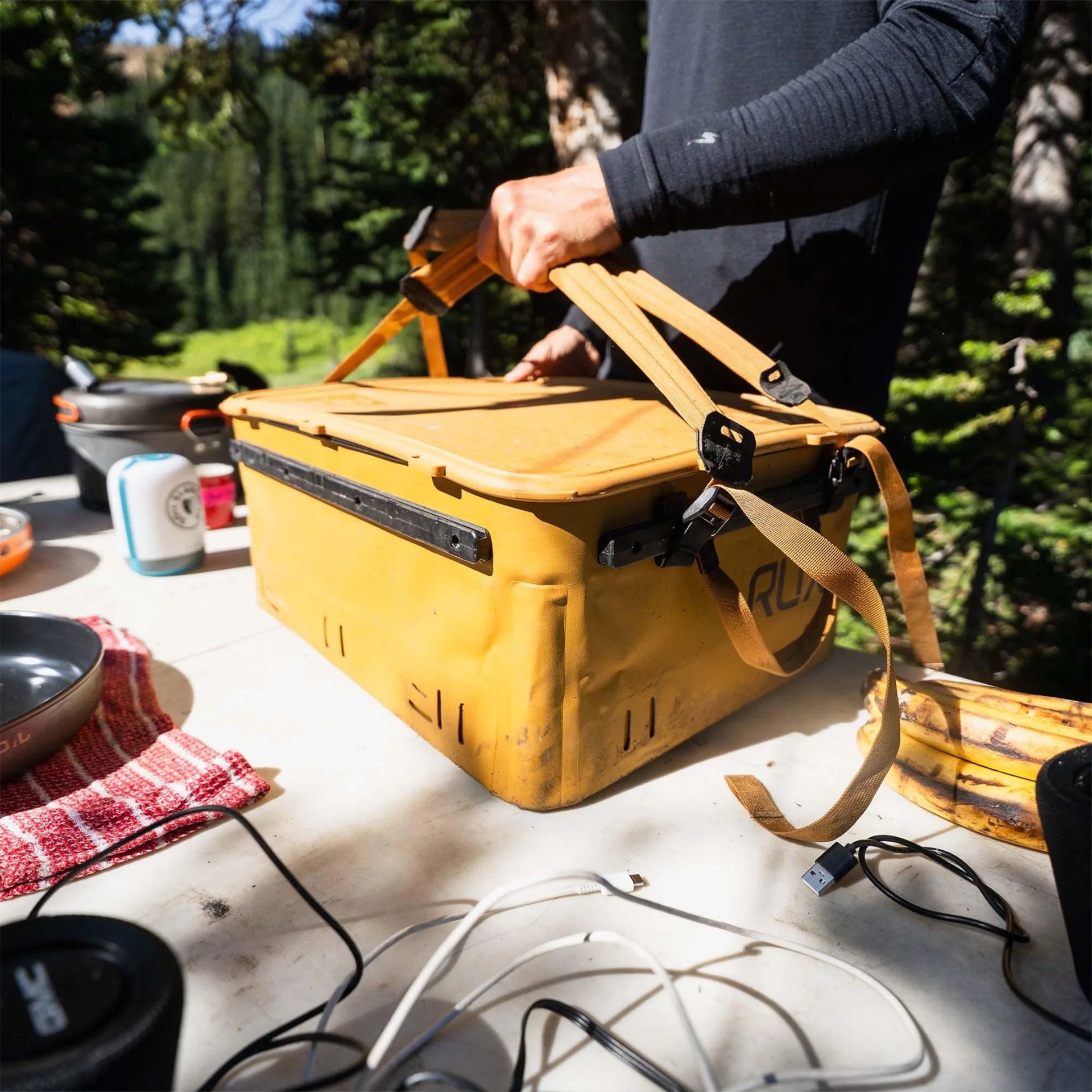 A person wearing a long-sleeve black shirt is holding the handles of a large, yellow, rugged cooler or storage box on a white outdoor table. The table has various items on it, including a red and white striped cloth, a black pan, some cables, a bunch of bananas, and other camping or outdoor gear. The background shows a forested area with green trees and sunlight filtering through.