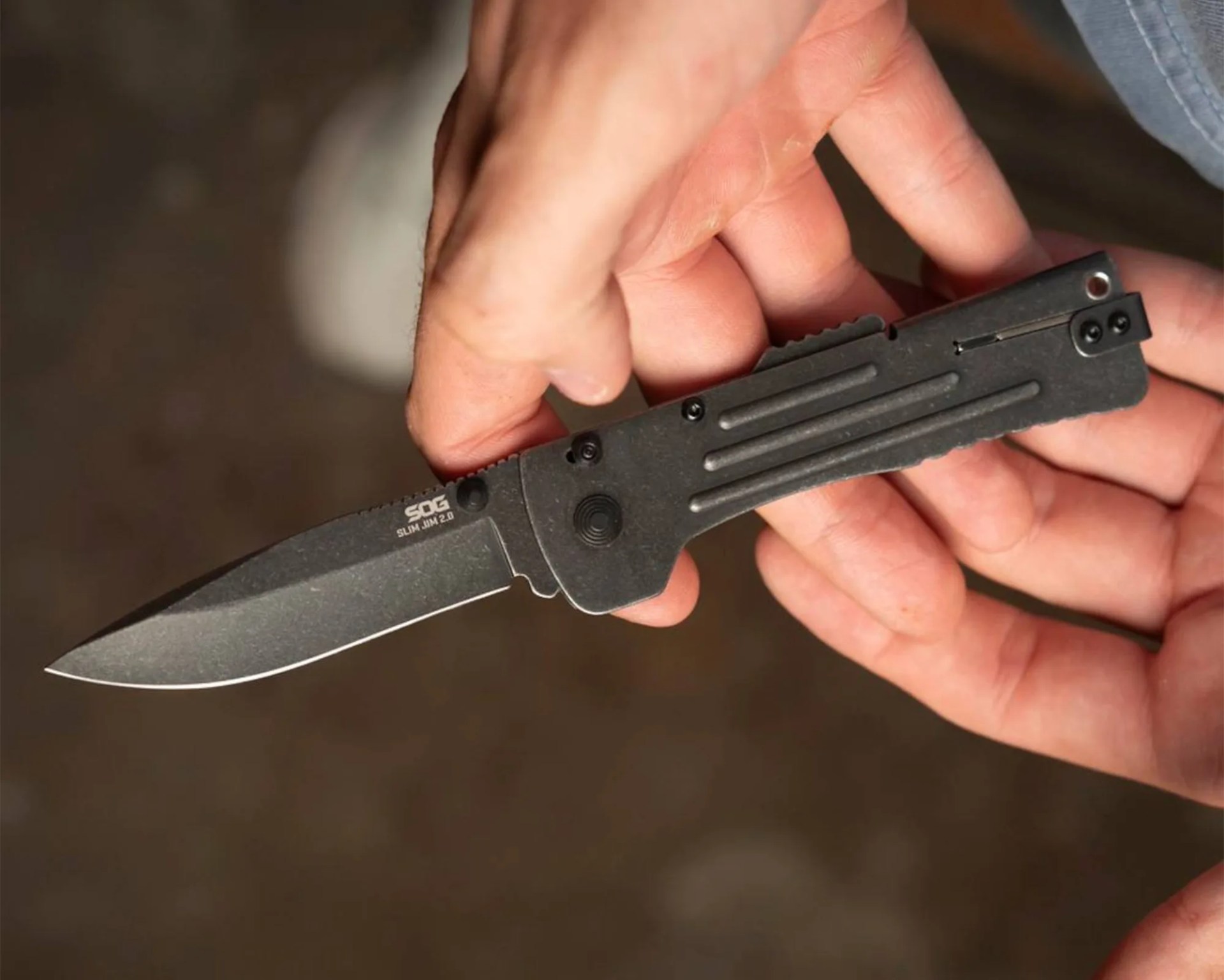 A hand holding a black folding knife with a partially serrated blade. The blade is marked with the brand "SOG" and the model "SLIM JIM 2.0." The handle has a textured grip and a pocket clip.