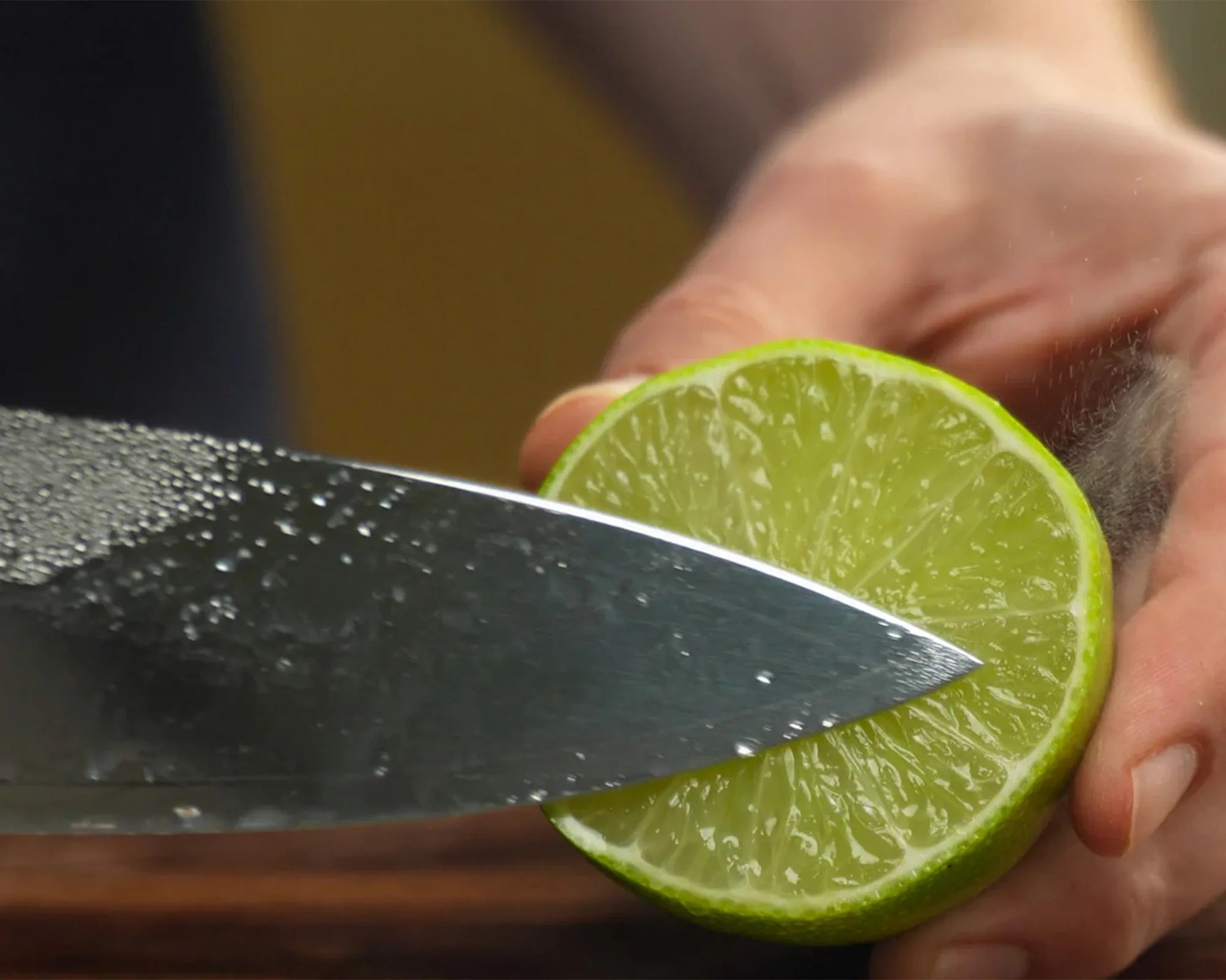 A close-up of a hand holding a halved lime while a knife presses against the lime's cut surface, with droplets of moisture visible on the knife blade.