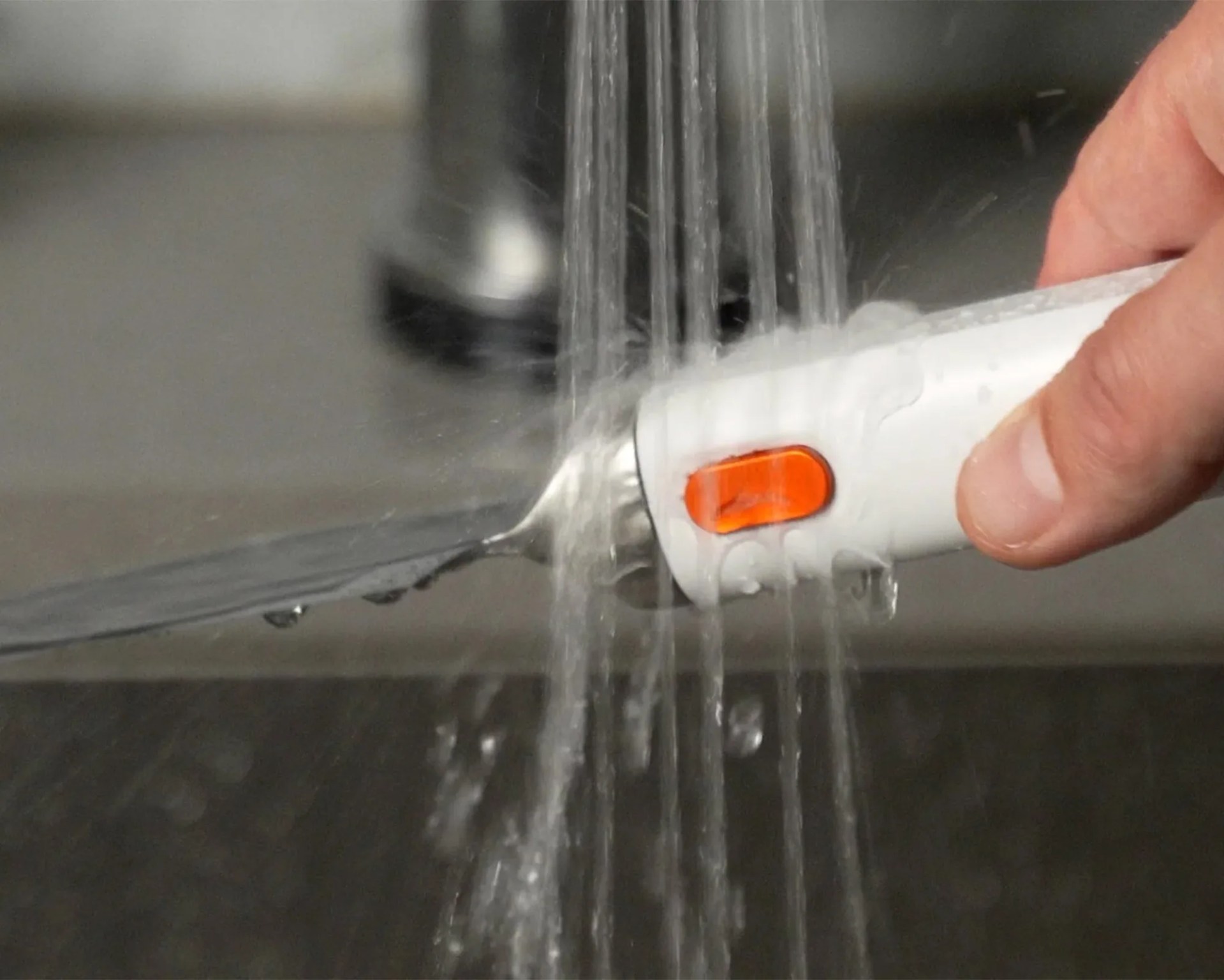 A hand holding a white electric knife with an orange button under running water, rinsing the blade.
