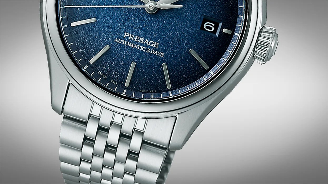 Close-up of a silver wristwatch with a textured dark blue dial, silver hour markers, and hands. The dial features the text "PRESAGE AUTOMATIC 3 DAYS" and a date window showing the number 6. The watch has a stainless steel bracelet and a ridged crown on the right side.