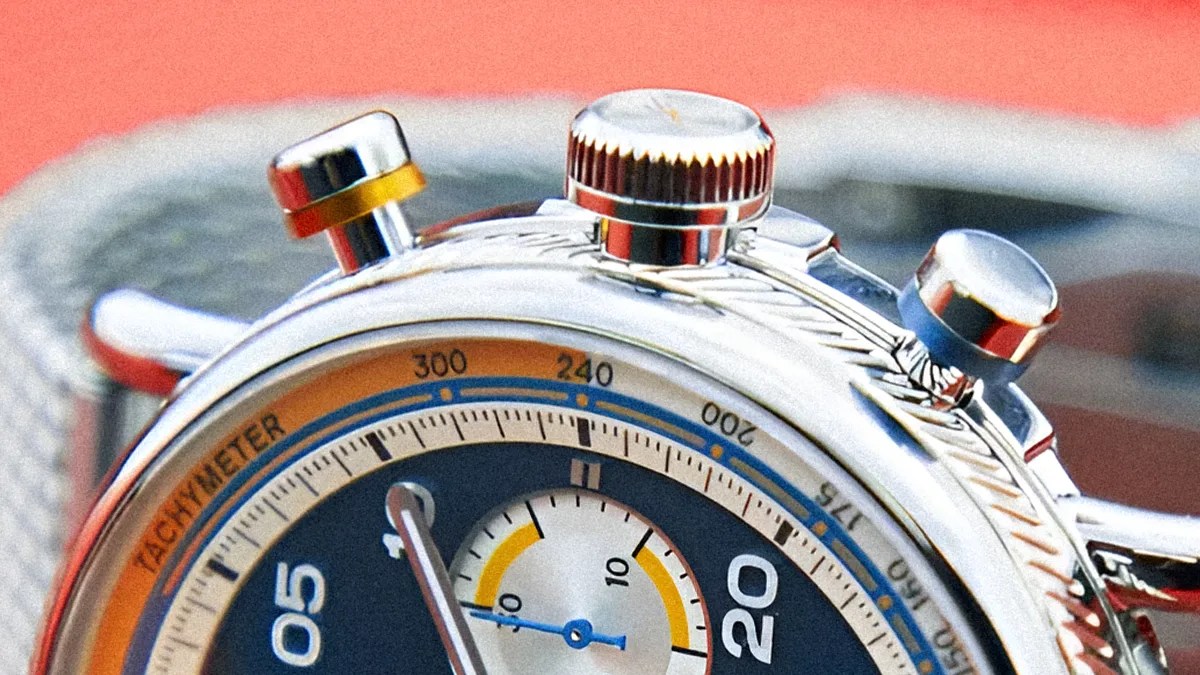 Close-up of a silver chronograph watch with a tachymeter scale on the bezel. The watch face includes a smaller sub-dial with yellow and black markings, and the main dial shows numbers 05 and 20 prominently. The watch has three push buttons on the side, one with a yellow ring and another with a blue ring. The background is blurred with a reddish hue.