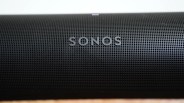 Close-up of a black Sonos speaker grille with the SONOS logo centered on the front. The grille has a pattern of small circular holes.