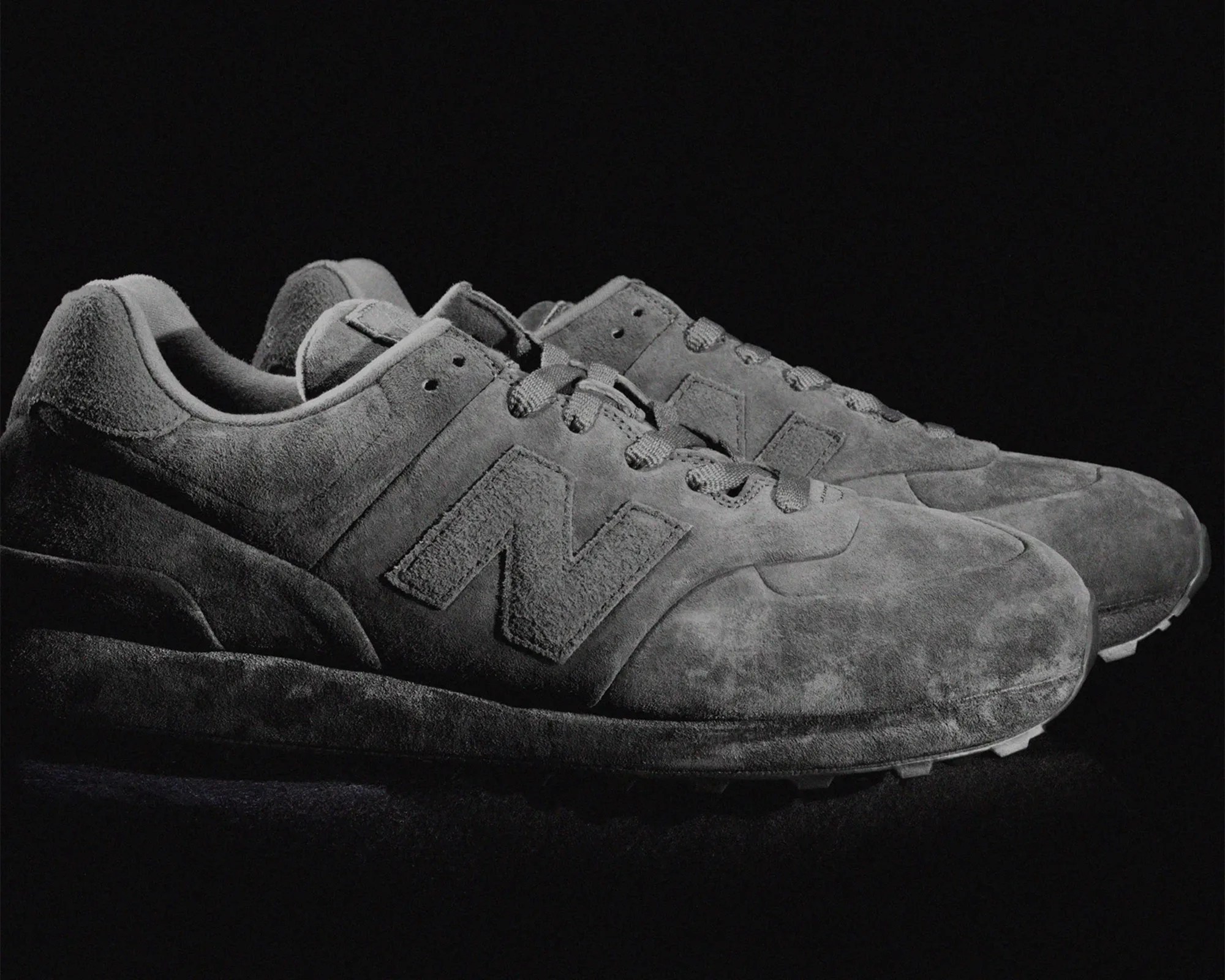 A pair of gray suede New Balance sneakers with the signature "N" logo on the side, displayed against a black background. The shoes have a textured, soft appearance with matching gray laces and a cushioned sole.