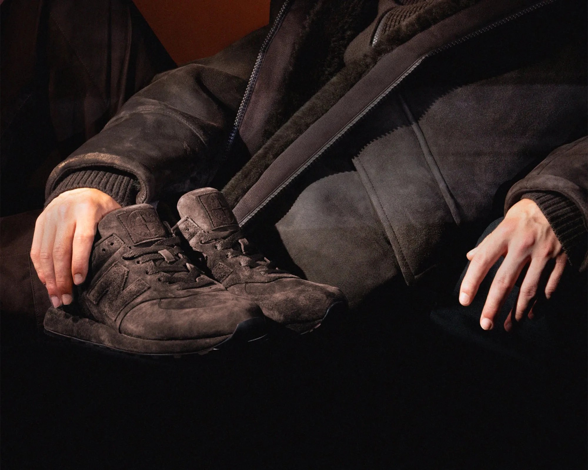A pair of dark brown suede sneakers held by a person wearing a matching dark brown suede jacket with a shearling lining. The sneakers have laces and a subtle embossed logo on the tongue. The jacket features ribbed cuffs and a zipper. The lighting highlights the texture of the suede material on both the shoes and the jacket.