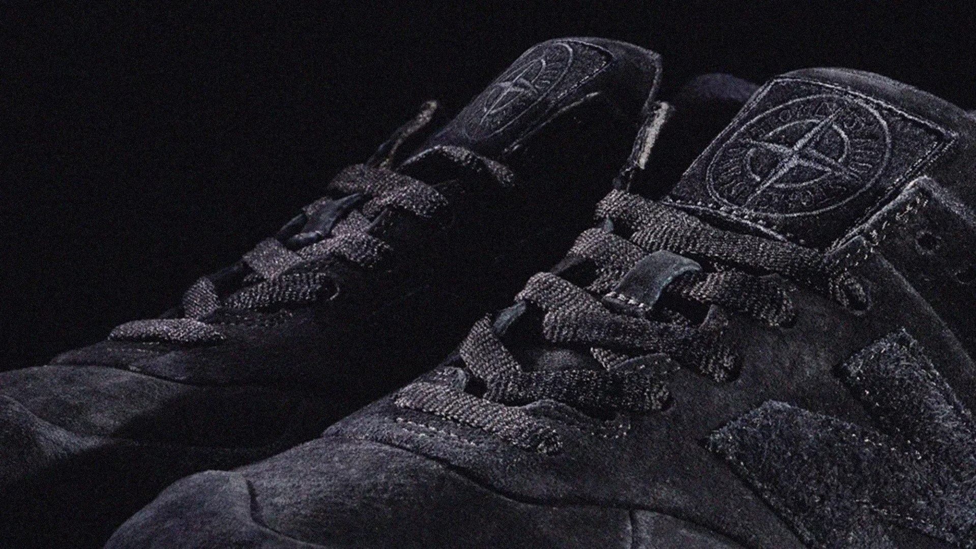 Close-up of a pair of black suede sneakers with black laces, featuring a circular emblem with a compass design on the tongue. The shoes have a textured "N" logo on the side. The background is dark, highlighting the shoes.