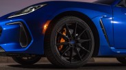 Close-up of the front left wheel of a blue Subaru BRZ, featuring a black multi-spoke rim, orange Brembo brake caliper, Michelin Pilot Sport tire, and a lit amber side marker light. The car's sleek blue fender and part of the headlight are also visible.