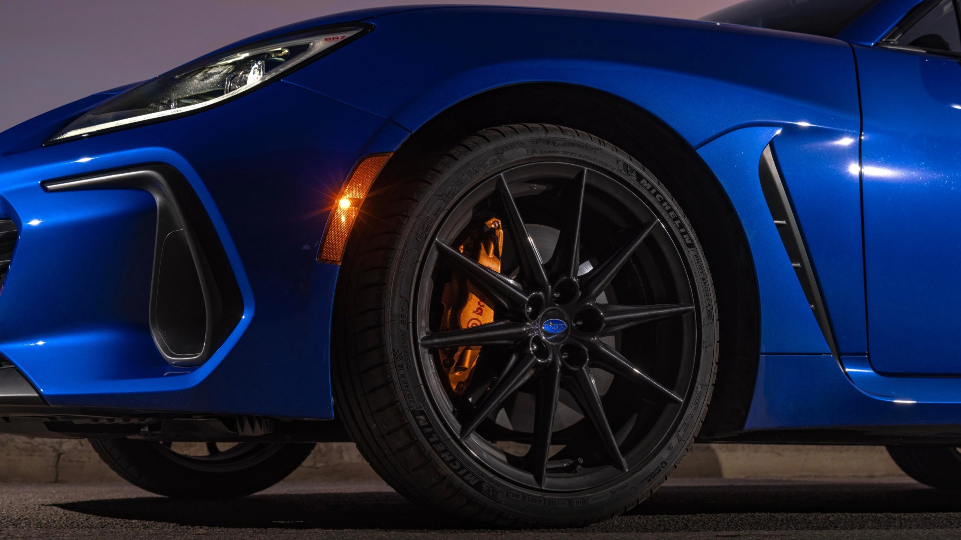Close-up of the front left wheel of a blue Subaru BRZ, featuring a black multi-spoke rim, orange Brembo brake caliper, Michelin Pilot Sport tire, and a lit amber side marker light. The car's sleek blue fender and part of the headlight are also visible.