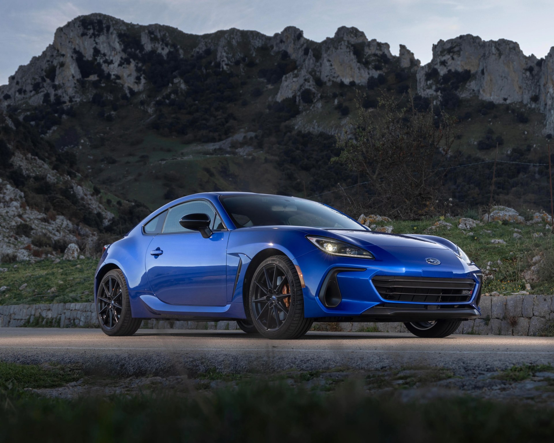 Blue Subaru BRZ sports car parked on a road with mountainous terrain in the background, featuring sleek aerodynamic design, black alloy wheels, and illuminated headlights.