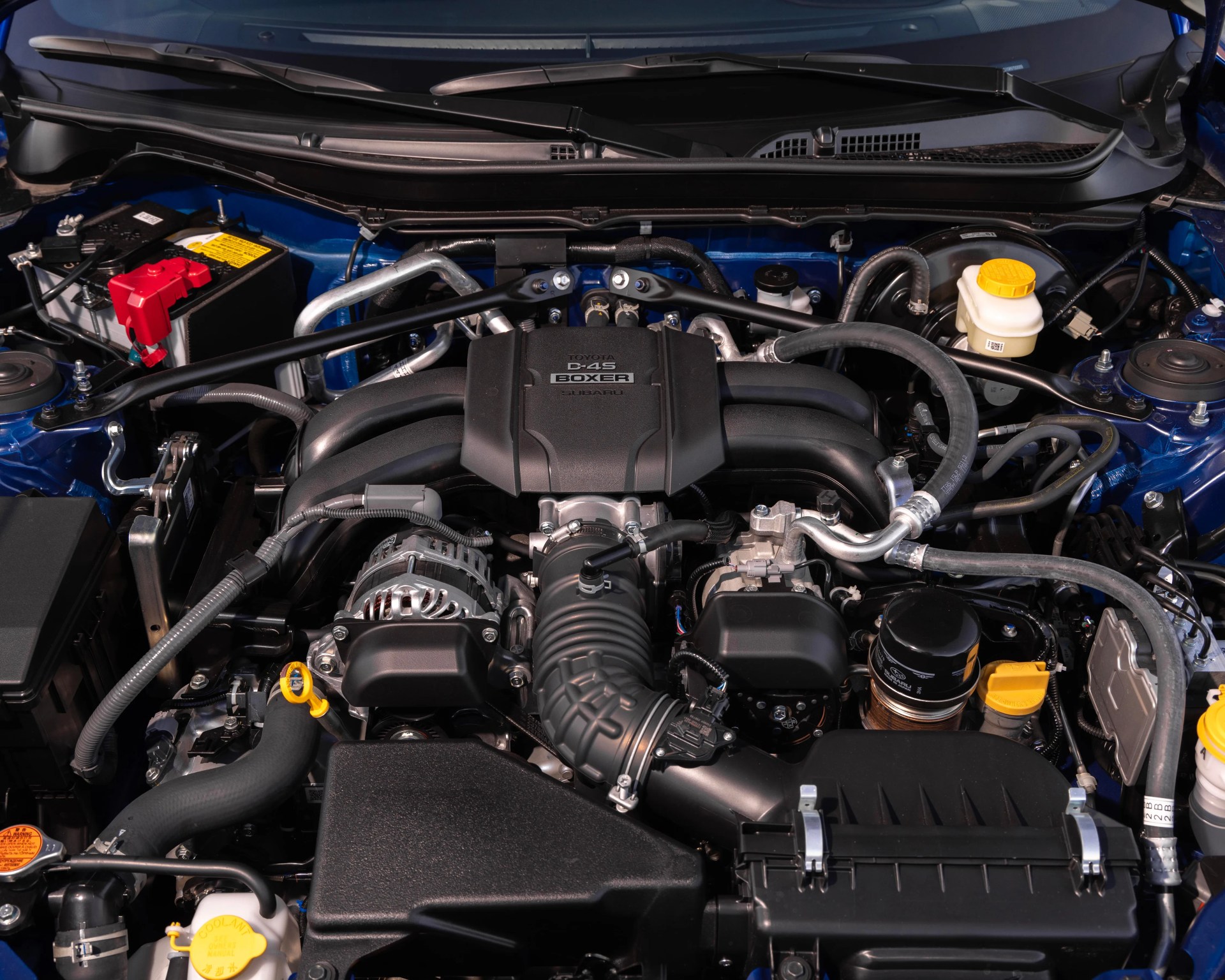Engine bay of a car featuring a Toyota D-4S Boxer engine co-developed with Subaru. The engine cover is black with "Toyota D-4S Boxer Subaru" branding. Visible components include various hoses, a battery with a red terminal cover on the left, fluid reservoirs with yellow caps, and an air intake system. The surrounding area is painted blue.